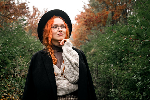 Fashionable women's autumn portrait in the park area. Red haired girl in a glasses, black hat with pigtails in a black coat stands against the background of withering foliage.