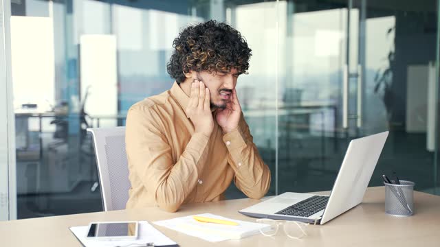 Sad employee suffering from toothache working on laptop while sitting at workplace at desk in modern office
