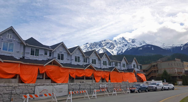 Construction Of New Modern Residential Housing In Small Town Pemberton British Columbia Pemberton, British Columbia, Canada- April 26,2023: Row of attached condos under construction. Pemberton BC.  Local landmark Mt Currie in background.  Draped in orange color tarp. Stone wall and row of Barricades in foreground.  Construction Paper covering stage. pemberton town stock pictures, royalty-free photos & images