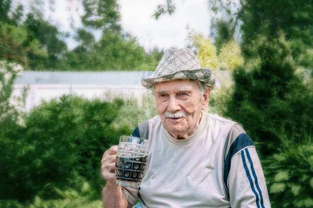 暗い飲み物のグ��ラスと帽子をかぶった白髪の老人の大きな肖像画。ガラスのマグカップを持った男が庭に座っている。 - beer glass mustache beer color image ストックフォトと画像