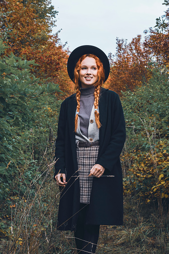 Fashionable women's autumn portrait in the park area. Red haired girl in a black hat with pigtails in a black coat stands against the background of withering foliage.