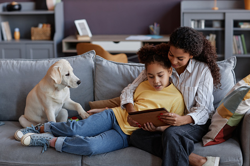 Full length portrait of loving mother and daughter using digital tablet together with cute dog while relaxing on sofa at home