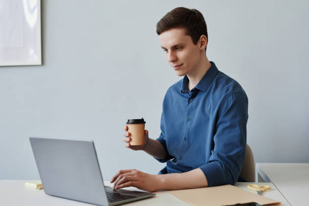 istock Smiling young man using laptop at workplace in office and enjoying cup of coffee 1499253580