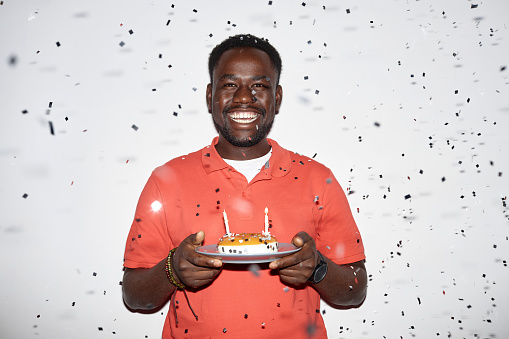 Minimal waist up portrait of smiling black man holding Birthday cake with confetti shower at party, shot with flash