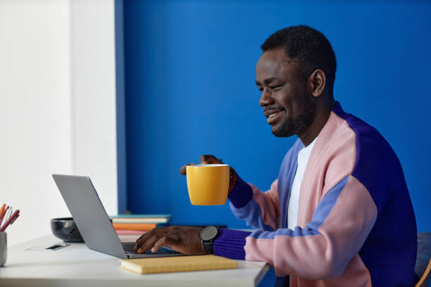 Black man using laptop at workplace in office with clock on blue wall