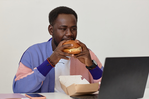 Portrait of young black man eating takeout burger at workplace in office against white wall, copy space