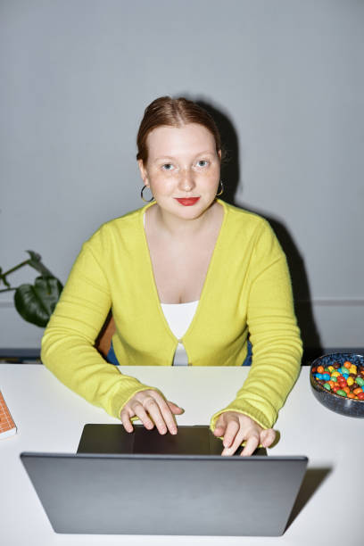 Red haired young woman using laptop at workplace and smiling at camera Vertical portrait of red haired young woman using laptop at workplace and smiling at camera shot with flash inside of flash stock pictures, royalty-free photos & images