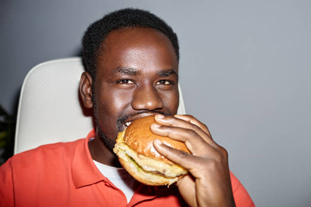 Closeup black man eating burger indoors and looking at camera happily