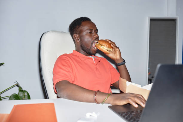 vista lateral homem negro comendo hambúrguer no local de trabalho e usando laptop - unhealthy eating flash - fotografias e filmes do acervo