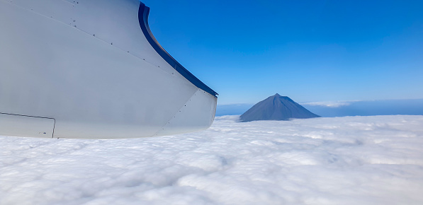 Aerial view from airplane window on Pico volcano above the clouds against blue sky, Azores.