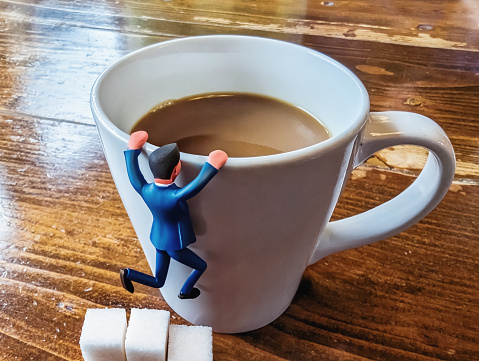 Photo of a generic toy businessman hanging on the side of a white ceramic cup of coffee, as if trying to climb in; 