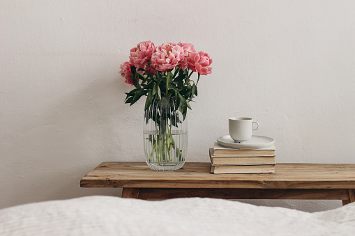 Feminine interior, still life. Rippled glass vase with coral pink peony flowers. Floral arrangement. Cup of coffee, books on vintage teak wooden bench, table. Empty white wall background. Blurred bed, Scandinavian bedroom.