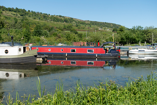 2nd June 2023 - Bowling, Scotland, UK: A tranquil scene of small boats, including a red and blue narrowboat, moored  at the side of the Forth and Clyde Canal at its western terminus at Bowling Harbour., West Dunbartonshire.
