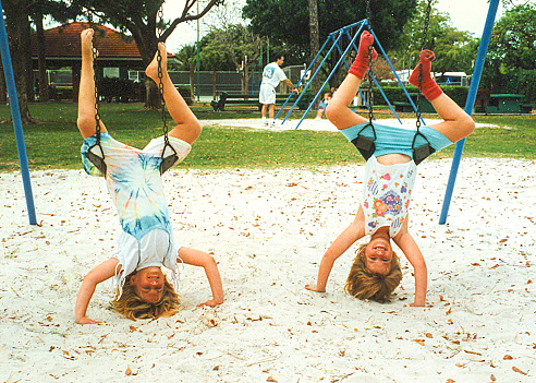 Twin girls are playing at the local playground. They are in a funny upside down position on the swings, looking toward the camera. It is bright shady daylight, no direct sunlight.