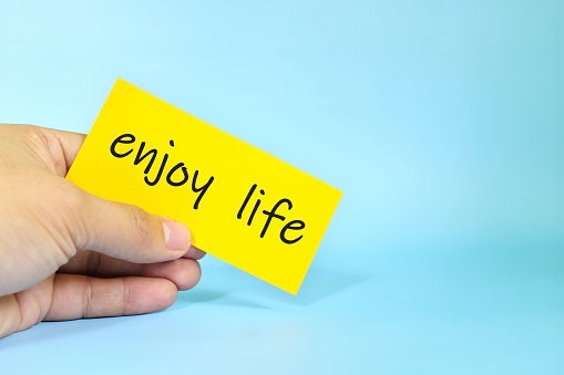 Enjoy life reminder concept. Hand holding a bright yellow paper message note.