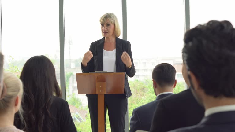 senior business woman standing at podium giving a speech addressing a conference in seminar. old executive female presentation in Meeting at office on window with colleagues laugh