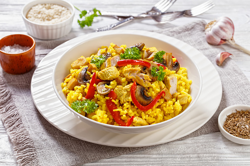 Chicken Paella of arborio rice, chicken fillet, mushrooms, julienned bell pepper and spices in white bowl on white wood table, spanish cuisine