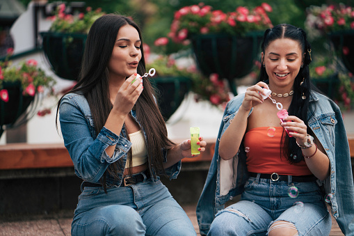 Two girlfriends sitting in park.  Girls sit and laugh, eat and have fun in the city park/center. They feel beautiful while the sun warms their summer days and awakens idyllic feelings in them. They sit in love and enjoy.