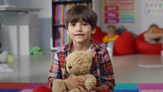 Cute preschool boy holding teddy bear and looking at camera in kindergarten. Portrait of adorable little kid playing with toy in playroom of nursery school