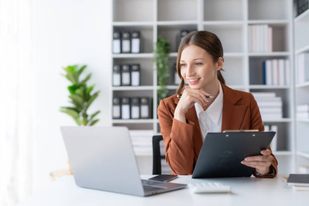 Beautiful young business woman manager or company worker holding accounting document, checking financial data or marketing report working in office with laptop. Accountant consults on some document. stock photo