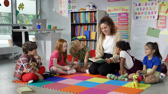 Female teacher reading story to group of elementary pupils in school classroom. Young teacher and preschool kids sit on classroom floor and read book together in kindergarten