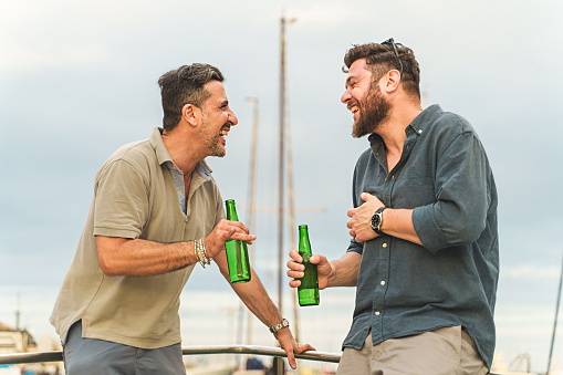 Two caucasian friends in their forties laughing heartily on the deck of a docked boat. They are wearing summer outfits and enjoying a joyful moment together holding beer bottles.