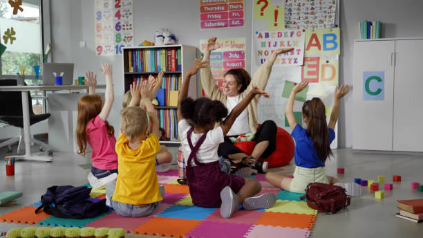 Elementary school teacher sit in class on floor with little pupils and moving hands up