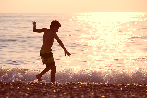 Silhouette of a boy by the sea at sunset. He runs along the shore, plays with the wave.