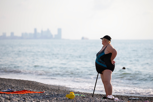 An elderly plump woman with a stick, a crutch and a fat belly is resting on the seashore.