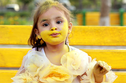 Cute baby girl of Indian ethnicity sitting in the public park outdoors and her both cheeks with yellow color powder paint on Holi festival. Holi is a popular and significant Hindu festival celebrated as the Festival of Colours, Love and Spring. It celebrates the eternal and divine love of the god Radha and Krishna.