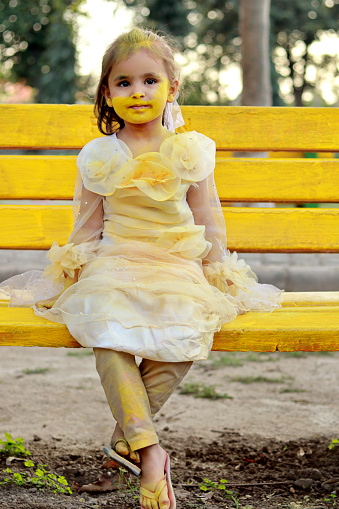 Cute baby girl of Indian ethnicity sitting in the public park outdoors and her both cheeks with yellow color powder paint on Holi festival. Holi is a popular and significant Hindu festival celebrated as the Festival of Colours, Love and Spring. It celebrates the eternal and divine love of the god Radha and Krishna.