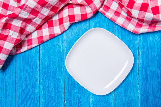 istock Top view background with squared empty white ceramic plate on vintage weathered blue wooden boards and red checkered kitchen towel on top. 1499390818