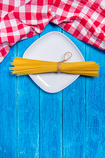 Top view background with squared white ceramic plate with spaghetti package on vintage weathered blue wooden boards and red checkered kitchen towel on top. Vertical photo