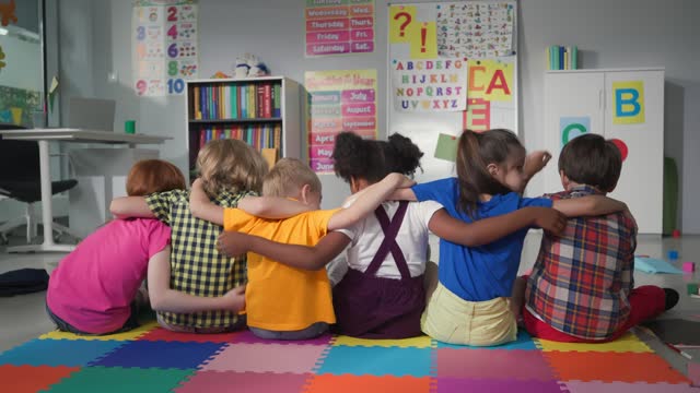 Back view of diverse preschool kids sit on floor in a row and hug in kindergarten