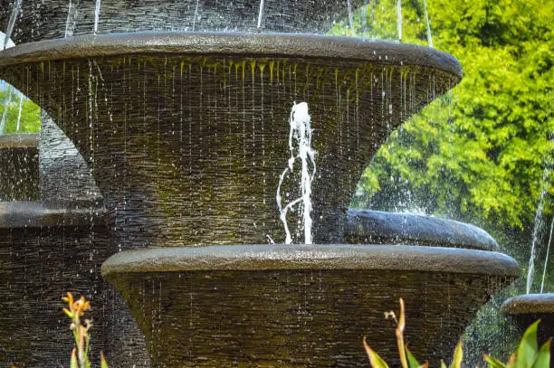 Photo of Close Up View Fresh Water Splash In The Multi-tiered Fountain Garden With Lush Leafy Trees Background