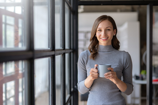 Successful businesswoman smiling holding coffee cup at office. Confident business woman standing drinking coffee in the office. Woman standing by the window.