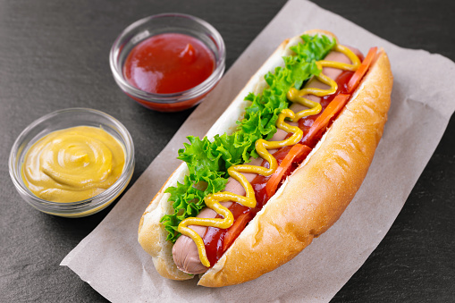 Homemade Hot Dog with yellow mustard, ketchup, tomato and fresh salad leaves on black slate background. Fast food, street food, american cuisine