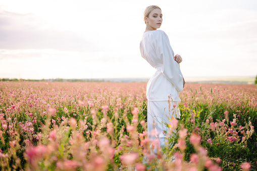 Young woman in stylish summer dress feeling free in the field with flowers in sunshine. Nature, vacation, relax and lifestyle. Summer landscape.