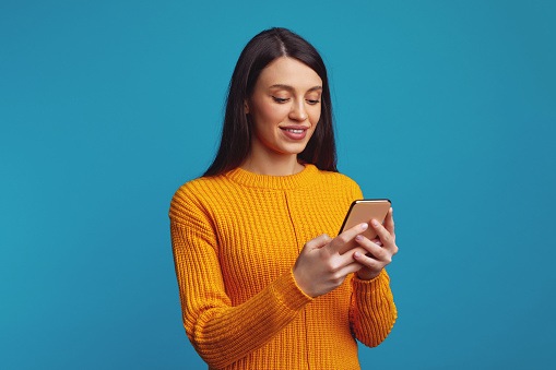 Smiling woman using smartphone, checks bank account or sends message in social networks, wears orange clothes, isolated over blue background