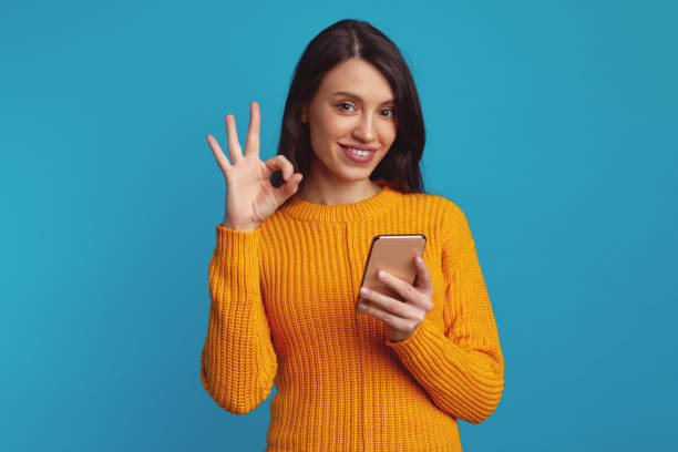 istock Young girl dressed in warm knitted orange sweater showing ok gesture while using smartphone 1499450080