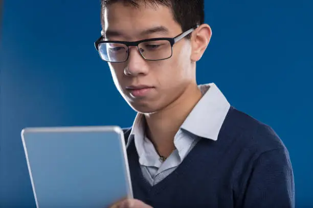 Photo of Engrossed Asian man checks tablet, blue backdrop