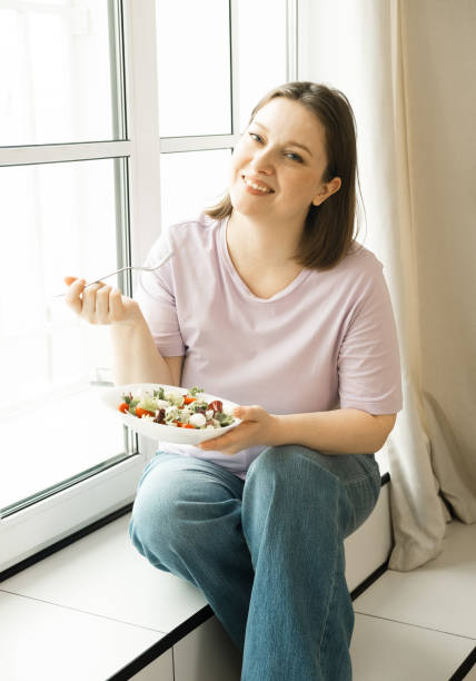istock Oversized woman next to window and eating fresh vegetable salad 1499458915