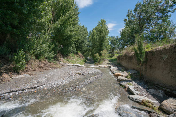 water flowing through a river in La Alpujarra de Granada, there are trees, there is a wall, there are stones and the sky has few clouds