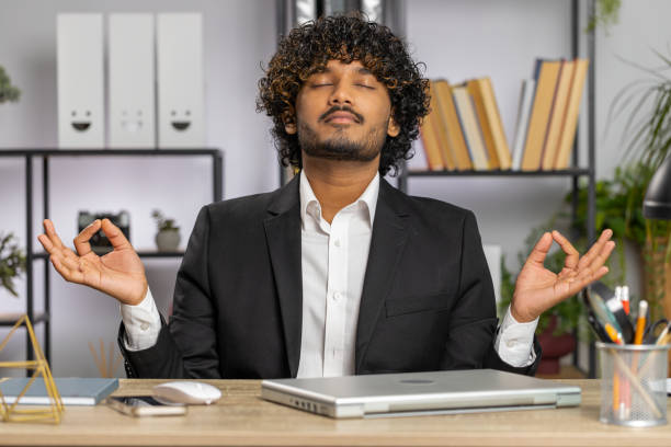 Businessman working on laptop, meditating, doing yoga breathing exercise in lotus position at office Indian businessman working on laptop computer, meditating, doing yoga breathing exercise in lotus position at home office. Calm serene freelancer arab man taking break. Busy occupation. Peace of mind meditation room office stock pictures, royalty-free photos & images