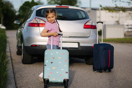 Adorable little girl standing in front of the car with her kid suitcase and waiting for parents. Family journey. Travel concept . High quality photo