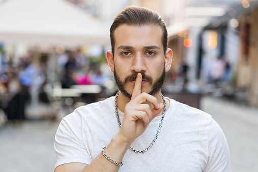 Shh be quiet please. Bearded young adult man presses index finger to lips makes silence hush gesture sign do not tells gossip secretoutdoors. Guy walking in urban city sunshine street. Town lifestyles