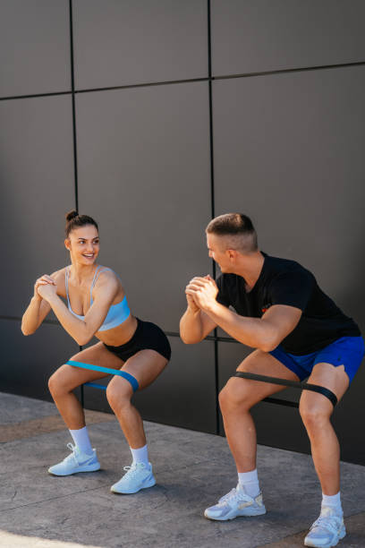 couple sportif faisant des squats avec des bandes de résistance enroulées autour de leurs jambes - crouching male men sensuality photos et images de collection