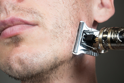 Man is cutting beard by Electric metal razor or dry shaver. Trimmer for cutting. Short, sparse beard on mans face. Hair growth problems. Unshaven bristles on the beard.