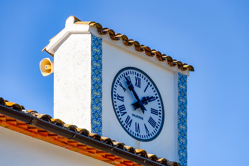 Guadalest, Alicante, Spain - January 14, 2023: A mini clock tower on a building's roof with Roman numerals showcasing the hour and minute hands. A public megaphone is visible behind the spire.