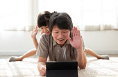 Happy Asian father and little girl lying cozy on the floor carpet chatting with grandparents by video conference on digital tablet, Waving hands, Saying Hello looking at screen in living room at home.